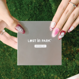 Model with medium almond Cherry Stardust press-on nails holding the LOST in PARK Artisan Set box on green grass.