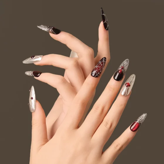 Artistic hand pose showcasing the “Crimson Whisper” nails with metallic silver designs, deep red shimmer, and white spiderweb graphics.