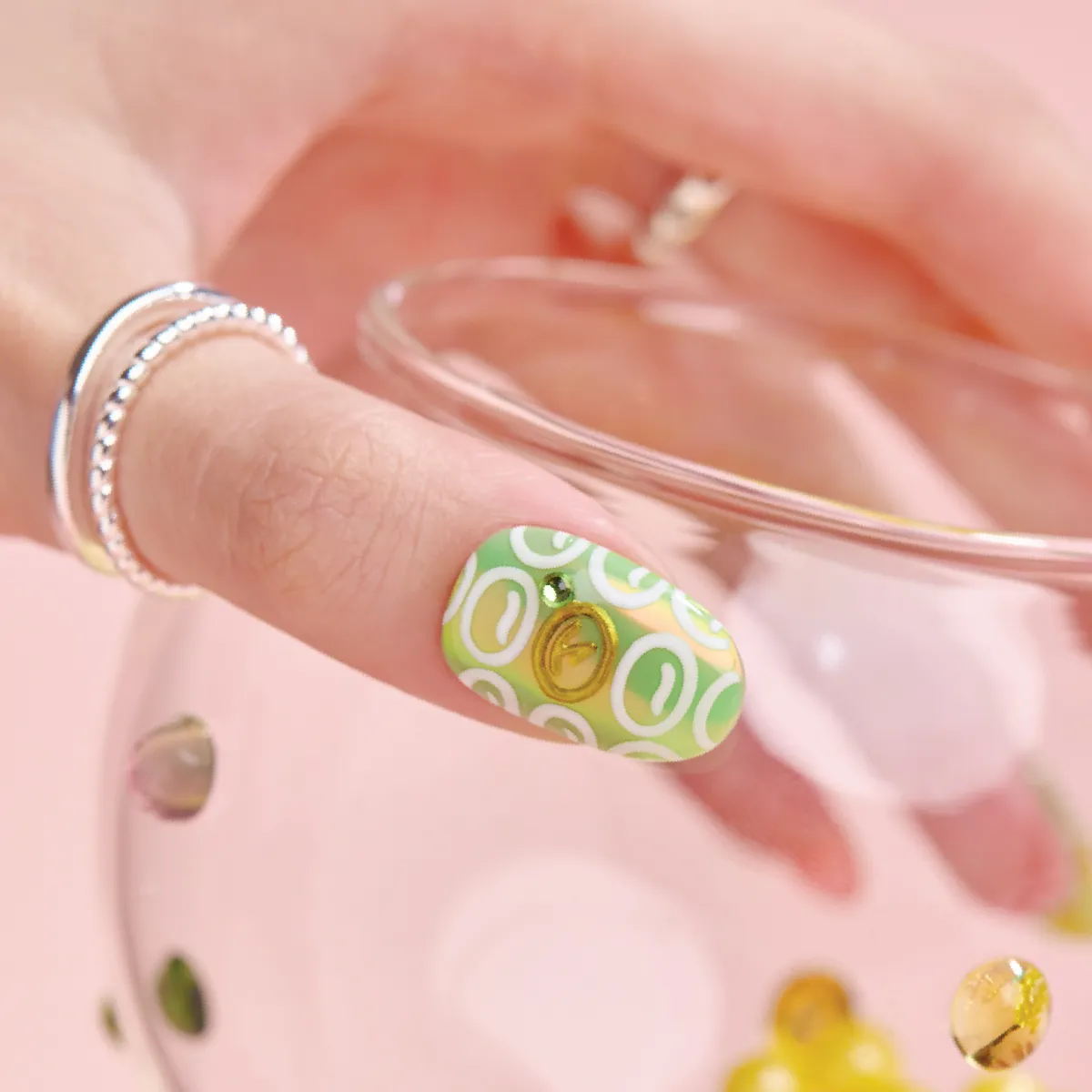 Close-up of a hand wearing a green press-on nail with retro-style white ovals and a gold embellishment, holding a glass bowl.
