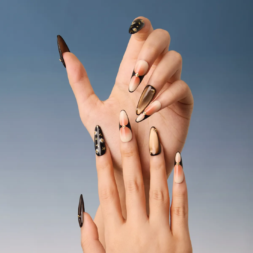 Hand model wearing long almond press-on nails in black, nude, and gold tones with gradient and gem details.