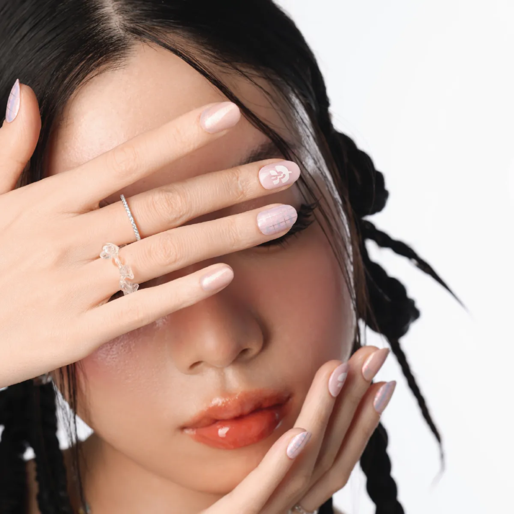Model with braided hair covering face with hand, wearing pink plaid press-on nails with white logo accent.